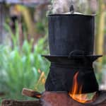 Black pot boiling for the rice cooker on the fired stove next to firewood pile.