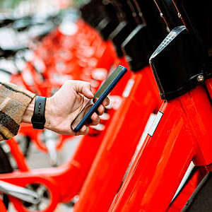 Close-up of a man’s hand using a smartphone to rent a red bicycle from an urban bicycle sharing station.