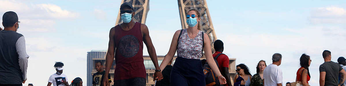 Tourist couple wearing protective masks walk along the Eiffel Tower esplanade in Paris holding hands.