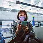 Young woman wearing a protective face mask taps on her smartphone as she waits for a night flight at a quiet airport terminal gate.